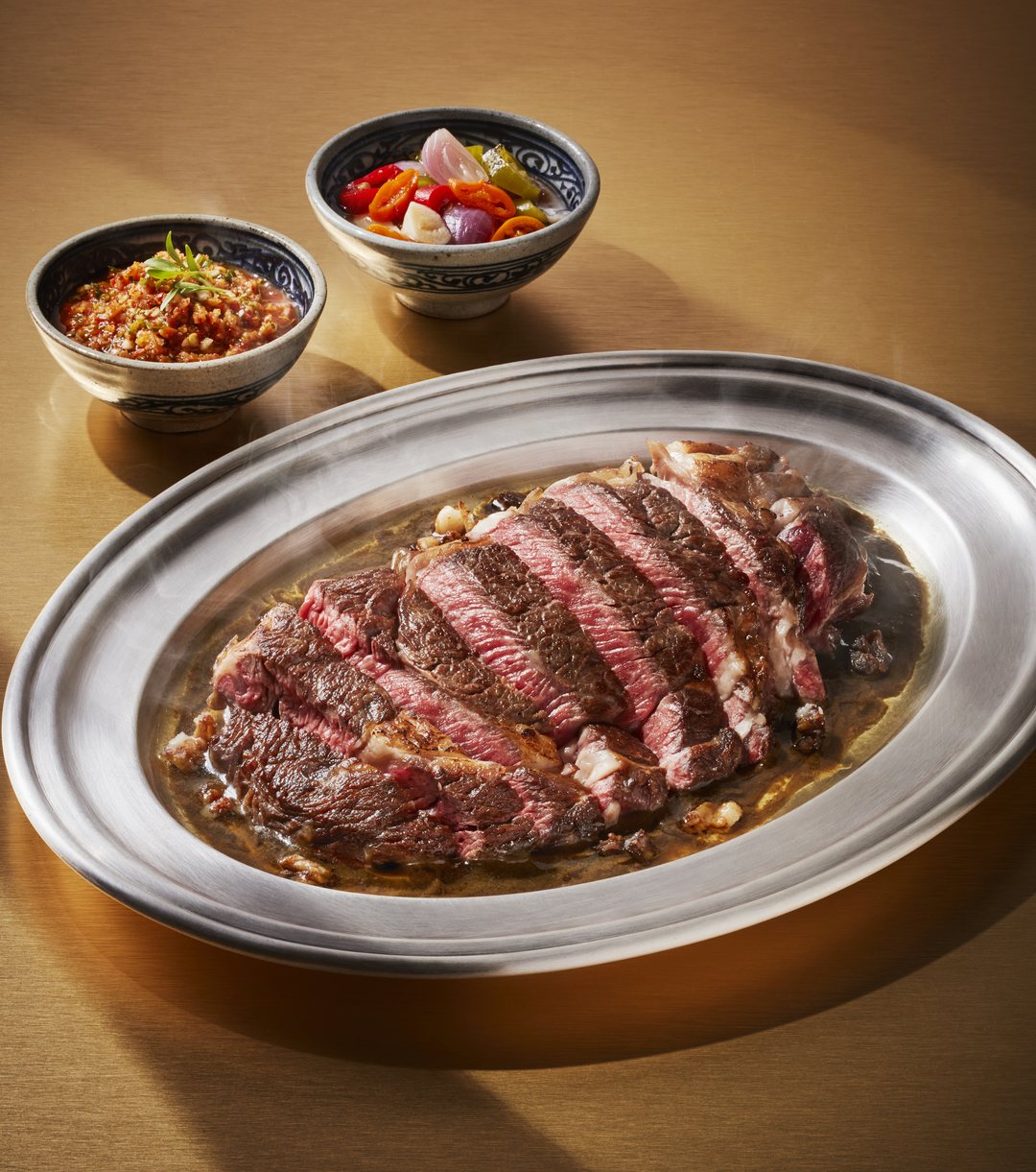 Sliced grilled ribeye on a silver platter, two small bowls of jaew behind it